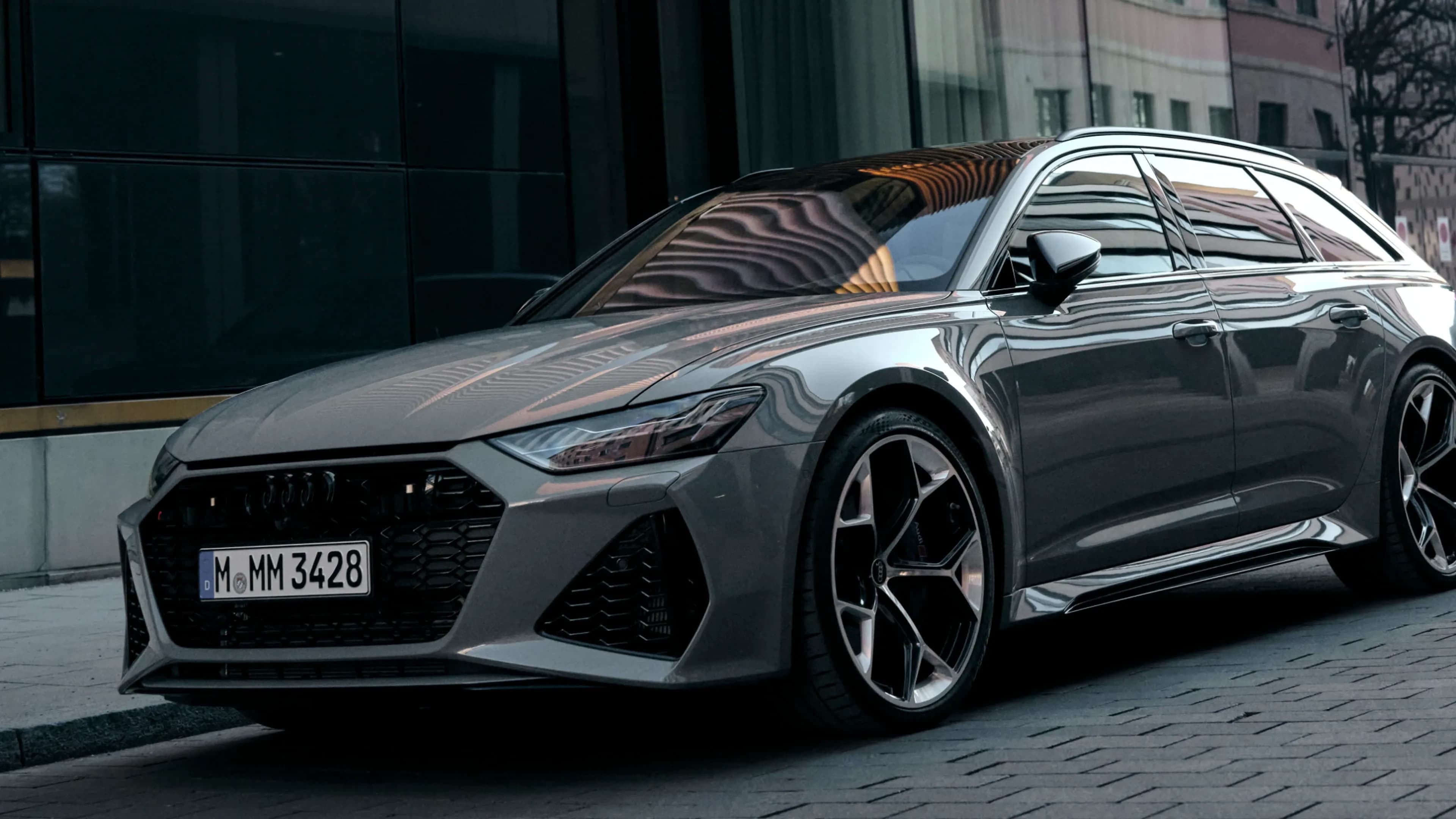 Grey Audi RS6 Avant in a low front three-quarter perspective on a Munich cobblestone street, available for luxury car rental at Mosaik Motors