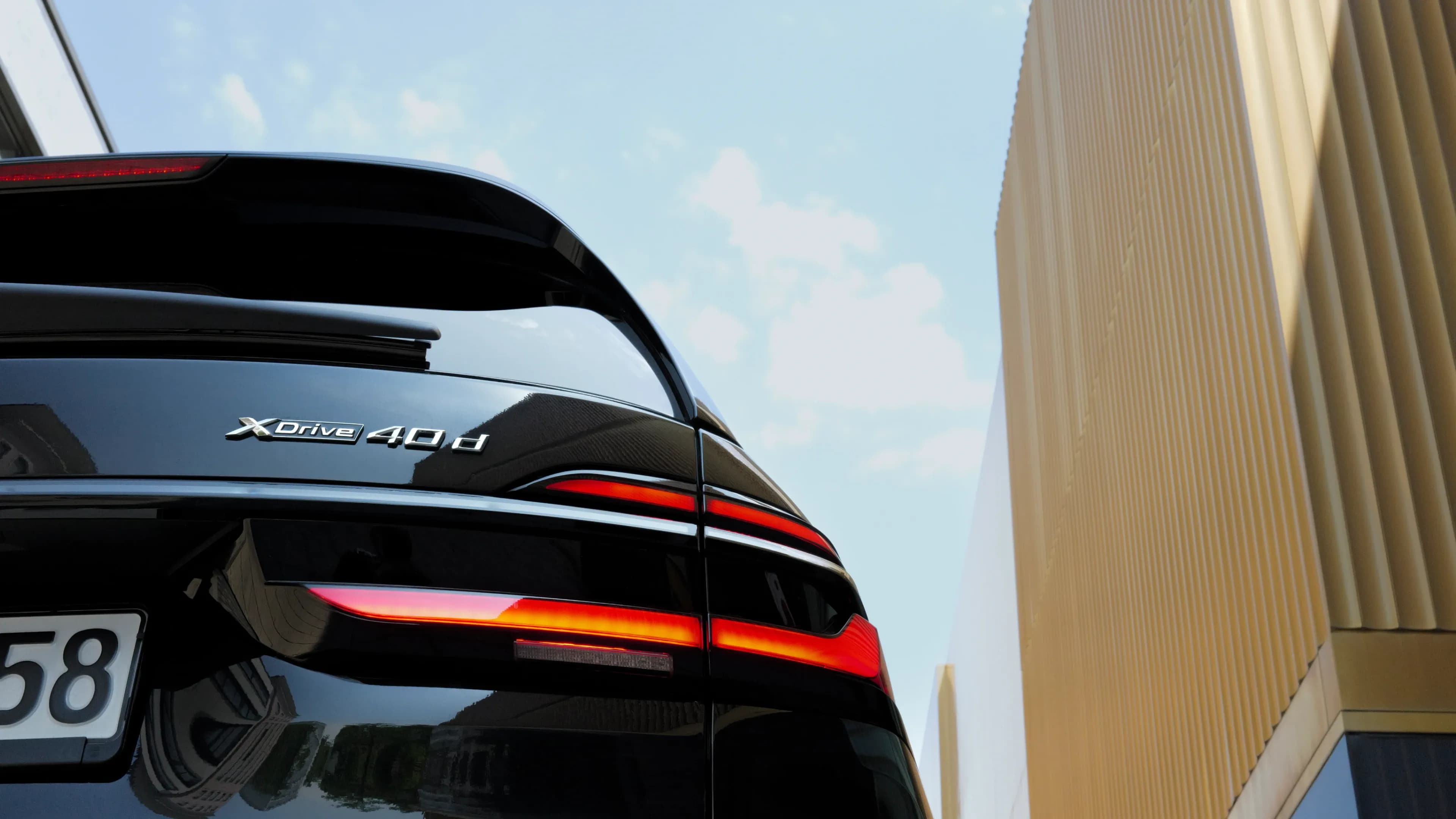 Close-up of the rear of a black BMW X7 xDrive40d showing the illuminated LED taillights in front of the modern architecture of the Lenbachhaus in Munich.