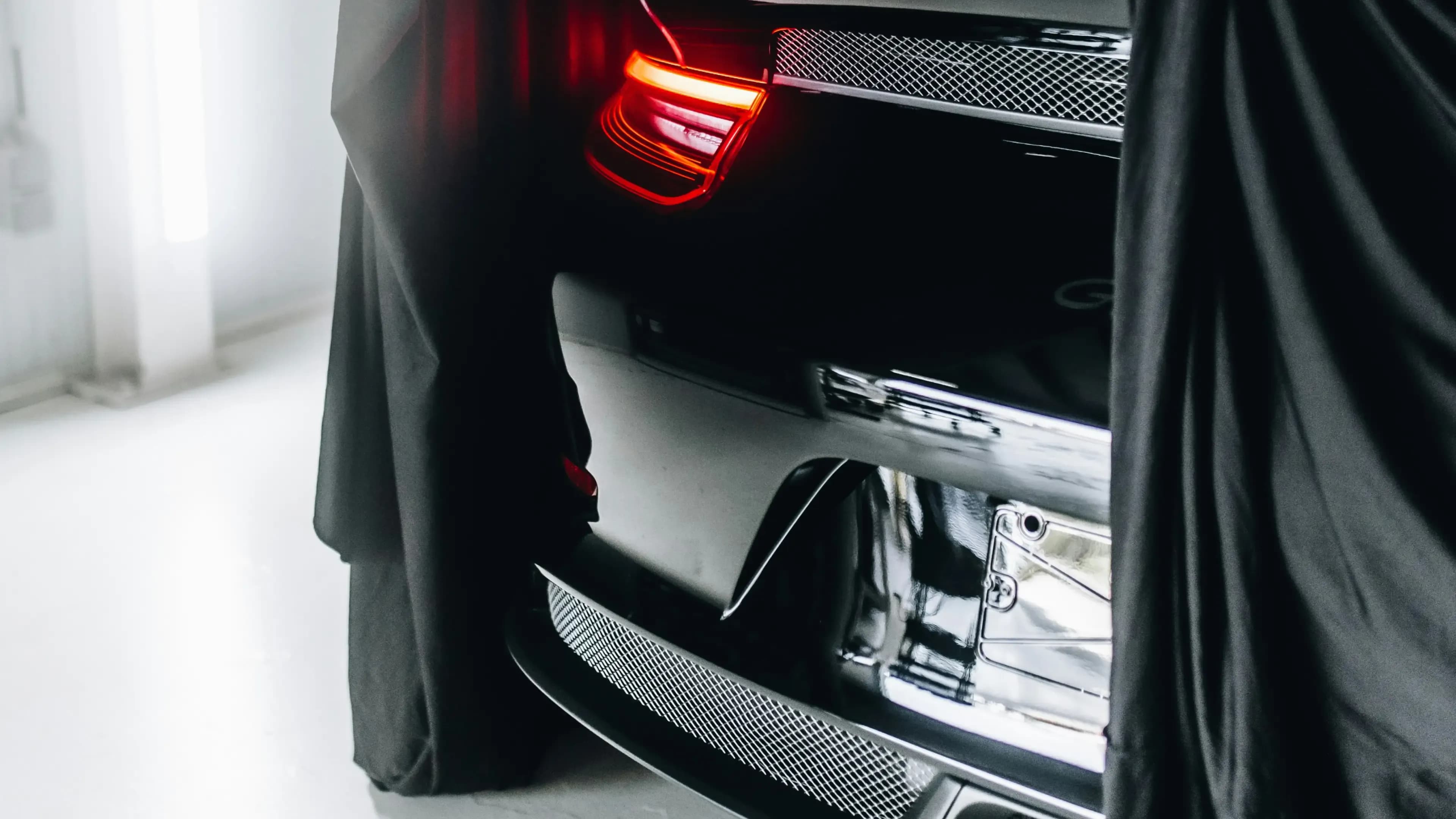 Mystery luxury vehicle with glowing tail lights partially revealed under black drape – Mosaik Motors bespoke special request service Munich